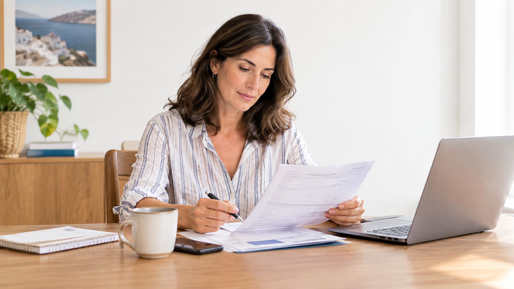 Mujer adulta revisando documentos financieros y facturas en una mesa de madera con su computadora portátil abierta.